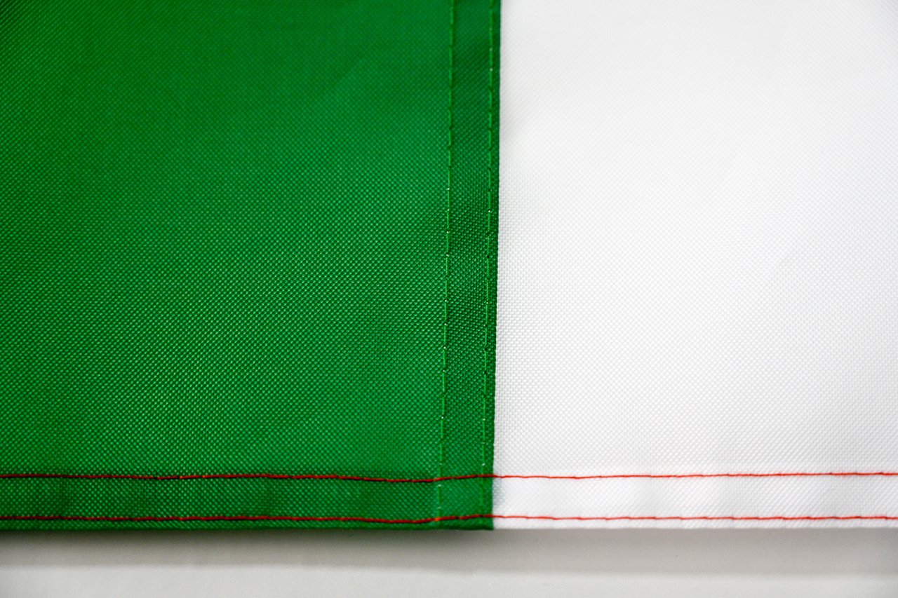 Flags-factory.com production line showing a sewing machine stitching the hem of a large 210D polyester flag, with workers inspecting the fabric